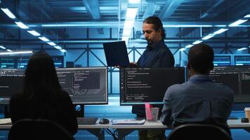 Middle eastern manager in server hub workplace doing inspection on electronics and multiracial workers. Team leader reading paperwork on laptop, verifying infrastructure documents, doing audit video