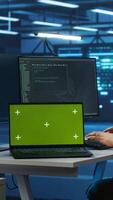 Vertical Engineer upgrading equipment in data center using green screen laptop. Specialist at PC desk in high tech establishment overseeing server rows with mockup notebook, panning shot video