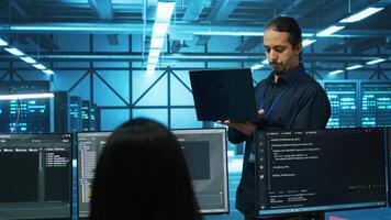 Engineer typing on laptop in server hub, checking recovery plan, monitoring data center energy consumption across components. Technician inspecting rackmounts, doing routine disk checks video