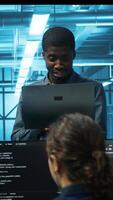 Vertical Smiling african american manager in data center doing checkup on electronics to avoid downtime. Team leader holding laptop supervising employees in server room infrastructure components video