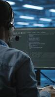 Vertical IT support worker in data center talking in headset mic with coworkers, coding on computer. Man discussing with colleagues using headphones microphone, overseeing server clusters video