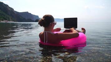 Woman freelancer works on laptop swimming in sea on pink inflatable ring. Happy tourist in sunglasses floating on inflatable donut and working on laptop computer in calm ocean. Remote working anywhere video