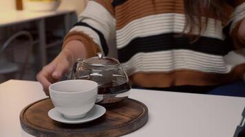 Cup of tea and kettle sitting on white kitchen table at home. Caucasian woman with sweater using tablet for call while drinking hot winter beverage from mug waiting for holiday dinner video