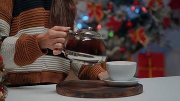 Close up of woman pouring tea from kettle in cup on kitchen counter at christmas prepared home. Person getting a hot drink in mug for cozy atmosphere while waiting on holiday celebration video