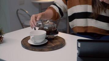 Close up of kettle and cup of tea on kitchen counter in christmas decorated home with tree. Happy woman pouring drink in mug while using tablet for call modern communication. video