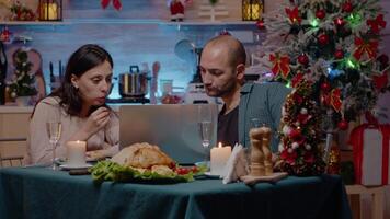 Couple looking at laptop and eating festive meal at christmas eve dinner. Cheerful people using device for entertainment while enjoying traditional food for holiday celebration. video
