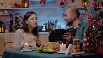 Couple enjoying christmas dinner and looking at tablet laughing and celebrating festive holiday at home. Man and woman watching device while clinking glasses with champagne and eating video