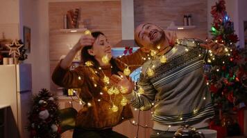 Cheerful couple getting tangled in string of twinkle lights for christmas eve preparations. Man and woman laughing while untangling knot of garland with lights and illuminated bulbs video