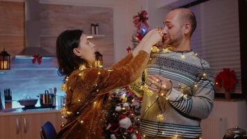 Festive couple trying to untangle garland of twinkle lights while decorating kitchen. Man and woman feeling cheerful and getting tangled in string of christmas lights with illuminated bulbs video