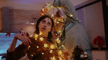 Man and woman tangled in garland of twinkle lights while decorating for christmas eve celebration. Couple laughing and untangling knot of string lights with illuminated bulbs. Cheerful people video