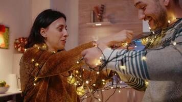 Couple laughing and getting tangled in string of christmas lights. Man and woman trying to untangle knot of garland with twinkle lights and illuminated bulbs. Holiday decorations video