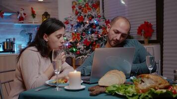 Festive couple looking at laptop and enjoying christmas dinner with technology. People eating chicken and drinking champagne while using device. Man and woman celebrating holiday. video