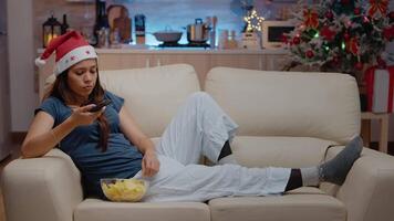 Bored woman switching channels and wearing santa hat on couch in festive space. Adult watching television and using TV remote control on christmas eve. Person with technology on holiday video