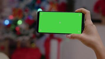 Close up of hand holding horizontal green screen on smartphone. Person looking at chroma key display with isolated template and mockup background in christmas decorated space video