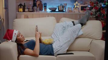 Woman browsing internet on smartphone and watching television on christmas eve. Festive adult looking at TV and phone screen for fun while eating chips from bowl. Person with santa hat video