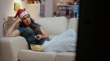 Bored adult holding TV remote control and switching channels on christmas eve. Lonely woman with santa hat looking at movie on television and having bowl with chips on couch. video