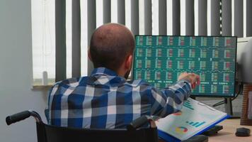 Handicapped businessman working with financial statistics checking graphs sitting immobilized in wheelchair on desk in building office. Paralysed freelacer using modern technology and documents. video