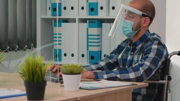 Handicapped businessman with visor and protection mask talking with colleague in financial company respecting social distance during global pandemic. Freelancer working in new normal business office. video