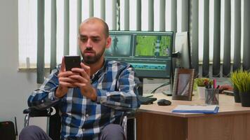Freelancer photo designer with disability in wheelchair texting on smartphone taking break from editing project creating content in modern company office. Blogger working from photo studio. video