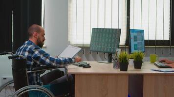 Handicapped paralysed businessman in wheelchair analysing economy data taking notes making financial expertise in business office discussing with colleague. Immobilized man using modern technology. video