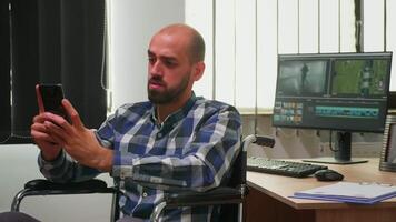 Disabled photo designer sitting in wheelchair searching on internet using smartphone texting, networking. Immobilized videographer editing new project working in modern company creating content video