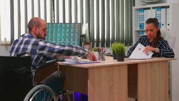 Businessman in wheelchair working in corporate office room changing documents analysing graphs. Employees in modern company typing on computer keyboard and pointing at desktop checking clients list. video