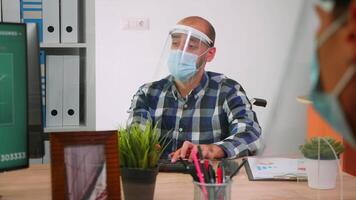Handicapped businessman with visor and protection mask discussing with colleague working in new normal business office. Man respecting social distance in financial company during global pandemic. video