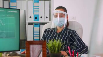 Company manager with face mask typing on computer and discussing with coworker during coronavirus pandemic in new normal business office respecting social distance. Freelancer using modern technology video