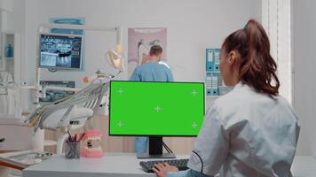 Woman using keyboard and computer with green screen in dental cabinet for oral care. Dentist working with chroma key on monitor with isolated mockup template for dentition and dentistry video