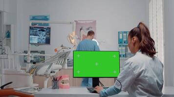 Dentist using green screen on monitor and talking to man assistant in oral care cabinet. Woman working with mockup template and chroma key on computer for teethcare and dentition video