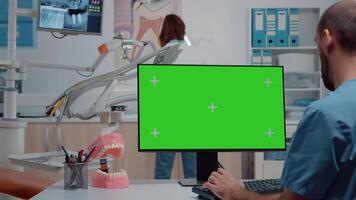 Man nurse looking at horizontal green screen on monitor in dental office while dentist working with dentistry equipment. Assistant using computer with chroma key and isolated template video