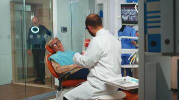 Young man dentist explaining the surgery using a plaster model of the mandible. Stomatologist holding mock-up of skeleton of teeth, sample of human jaw with toothbrush speaking with elderly patient. video