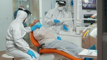 Orthodontist in protection coverall speaking to patient with toothache during covid-19 in stomatological office. Concept of new normal dentist visit in coronavirus outbreak wearing protective suit and face shield video