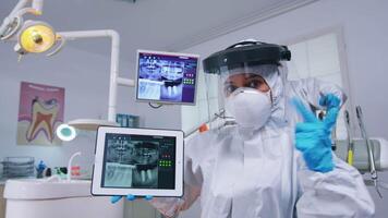 Patient pov of dental team explaining dental radiography and diagnosis for teeth infection using tablet. Stomatology specialist wearing protective hazmat suit against coroanvirus showing x-ray. video