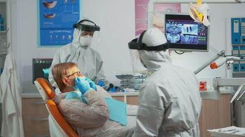 Senior woman sitting in dental clinic taking care of tooth health during covid-19 pandemic. Concept of new normal dentist visit in coronavirus outbreak wearing protective suit and face shield video