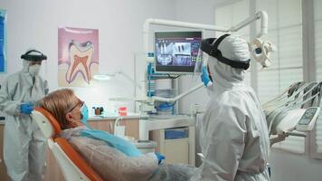 Orthodontist in special equipment pointing on digital x-ray explaining dental treatment during global pandemic. Medical team talking with woman wearing face shield, protection suit, mask and gloves video