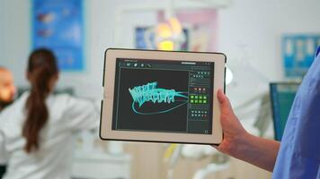Close up dentist assistant holding tablet with digital dental fingerprint of patient, while doctor is working with patient in background examining teeth problem sitting in modern dental clinic. video