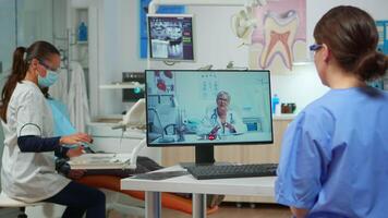Assistent having call with expert stomatologic dotor using computer while doctor is working with patient in background. Nurse listening dentist on webcam sitting on chair in stomatological office video