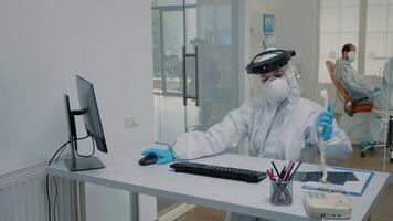 Dentistry nurse in protection suit sitting at desk using telephone and computer at dental clinic. Assistant working on appointments while stomatologist consulting patient in background video