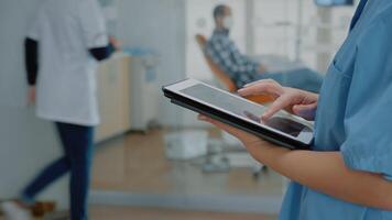 Close up of modern tablet with touchscreen and nurse hand using digital technology for development. Assistant holding device while stomatologist in cabinet consulting patient for oral care video