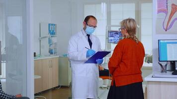 Orthodontist with mask speaking with elderly woman standing in waiting area of stomatological clinic taking notes on clipboard. Nurse typing on computer appointments in modern crowded office video