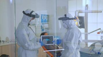 Dentist doctors in overall using tablet explaining dental x ray in stomatologic office during coronavirus. Man wearing face shield and mask showing to nusrse radiography using digital device video