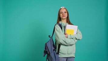 Portrait of joyous teenager with rucksack holding school notebook and notes, isolated over studio background. Jolly pupil with school supplies in arms, preparing to go to college, camera B video