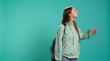 Curious smiling girl holding rucksack looking around for something, doing hand gesturing. Upbeat person doing surveillance gesture, exploring, seeking things, isolated over studio backdrop, camera B video