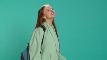 Joyous girl holding rucksack looking around, inviting others to join her, doing hand gesturing. Jolly teenager doing inspecting gesture, discovering new place, isolated over studio backdrop, camera A video