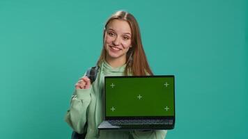 Smiling woman presenting laptop with green screen display, isolated over blue studio background. Cheerful person creating promotion with chroma key notebook device, camera A video