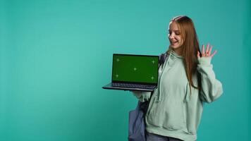 Happy girl presenting laptop with green screen display, isolated over studio backdrop. Radiant teenager creating promoting advertisement with mockup notebook device, camera B video