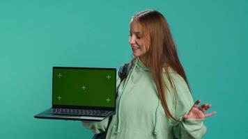 Happy girl presenting laptop with green screen display, isolated over studio backdrop. Radiant teenager creating promoting advertisement with mockup notebook device, camera A video