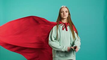 Happy young girl portraying superhero flying with cape fluttering in wind, isolated over studio background. Portrait of upbeat heroic woman posing as hero in costume, camera B video