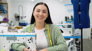 Female dressmaker working with sewing machine at textile factory and looking at camera video